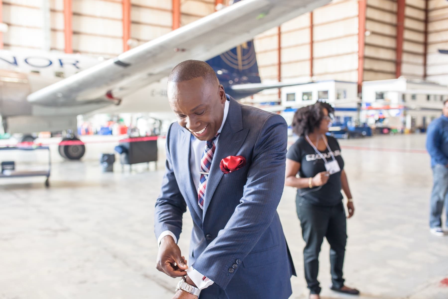 Model in a custom EZ Couture suit adjusting his cufflink inside a private-jet hangar, EZCOUTURE-branded crew shirt visible in the background
